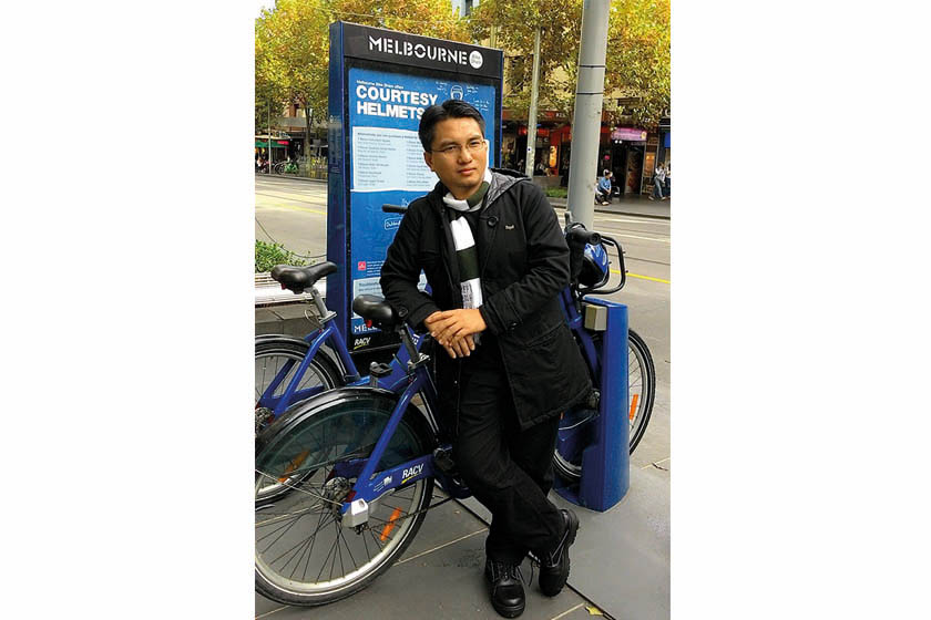 A man with glasses wearing black coat and striped shawl is reclining in front of the blue bike